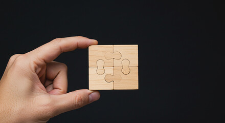 A hand holding a small square puzzle made of four wooden pieces against a dark background in a studio