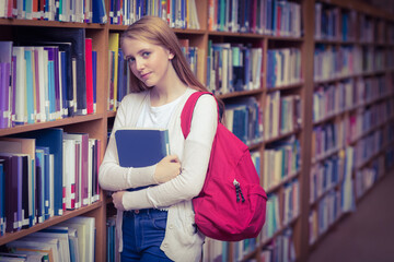 Leaning teenage girl holding blue book in library aisle, with red backpack and wooden shelves