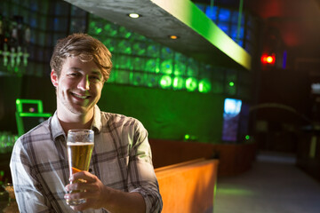 Smiling man standing at dimly lit pub bar counter, with tall beer glass, copy space