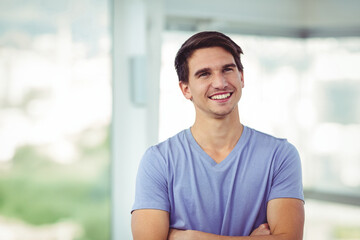 Fototapeta premium Smiling male in mid-20s standing with arms crossed in modern interior, featuring blurred cityscape