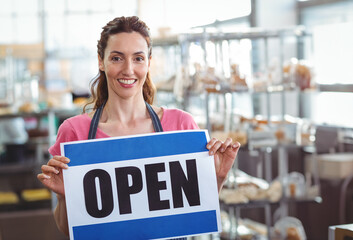 Smiling woman baker holding OPEN sign inside bakery, with metal racks and pastries