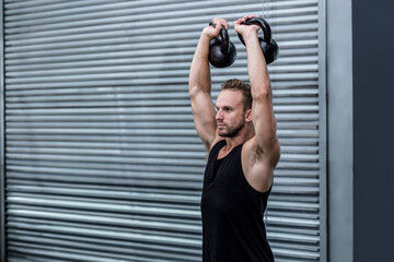 Lifting athletic man pressing two kettlebells overhead before garage door, with industrial theme