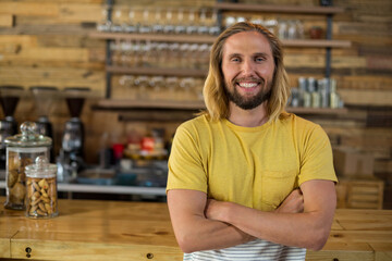 Fototapeta premium Smiling male barista standing behind coffee shop counter, with glass jars and espresso machine