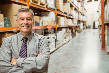 Fototapeta premium Standing middle-aged man with crossed arms in warehouse aisle with shelving and boxes, copy space