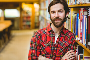 Obraz premium Smiling man leaning against bookshelf in library aisle, with rows of books and tables and chairs