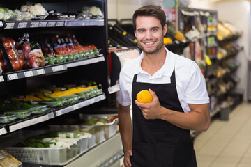 Smiling male grocery store worker holding orange in produce aisle, with packaged fruits, copy space