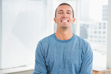 Smiling man wearing blue sweater standing in modern office studio, with blinds and city skyline