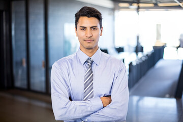 Fototapeta premium Standing Indian man crossing arms in modern open-plan office, with striped tie and glass partitions
