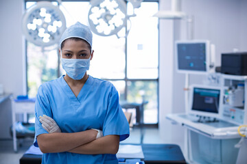 Standing woman surgeon crossing arms, gazing ahead in hospital operating room, with surgical lights
