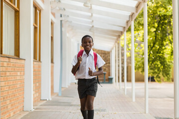 Obraz premium Walking African American school boy carrying red backpack along covered outdoor corridor, sunny day