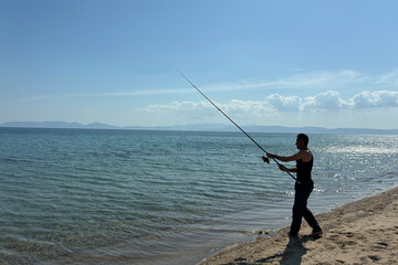 Man fishing on beach at sunset, catching fish rod into calm sea or ocean, outdoor adventure,...