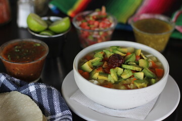 A vibrant bowl of traditional Mexican tlalpeño soup filled with fresh ingredients, avocado, and served with various condiments, perfect for a comforting meal experience.