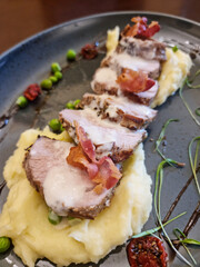 Danish - An elegant appetizer dish featuring sliced pork tenderloin on mashed potatoes, garnished with baked tomatoes, peas, and crispy bacon.