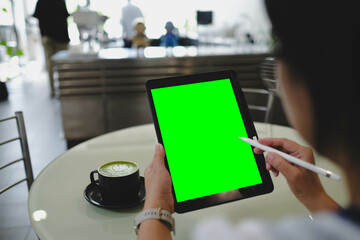 Person holding digital tablet with green screen and stylus at round table in coffee shop with matcha latte nearby.