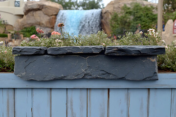 Fototapeta premium Stone planter box with flowers on a light blue wooden fence, waterfall in the background.