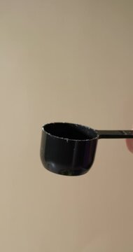 A close-up shot of protein powder gently tipping out of a black scoop held by hand, as fine particles fall in slow motion against a neutral beige background, suggesting vanilla or banana flavor.