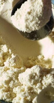 A black scoop presses into a fluffy, pale yellow protein powder, possibly vanilla or banana flavored, creating a soft texture detail in this close-up shot under warm, directional light.