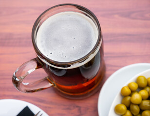 Glass mug of chilled dark beer with foamy head, served alongside plate of green pickled olives, creating contrast of textures and flavors, perfect for refreshing snack