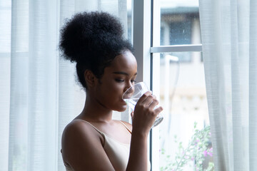 Beauty African woman enjoys healthy hydration, sipping clear water by a bright window; natural daylight reveals glowing skin and relaxed posture, reflecting modern wellness and mindful self-care