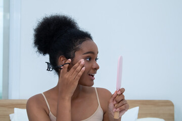 Beauty African woman with healthy skin curls lashes before pink mirror, smiling as she presses curler and checks result on bed; cozy evening light reveals complexion and playful self-care routine
