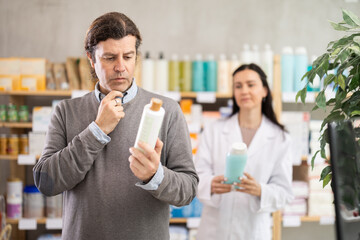 Attentive middle-aged man choosing shampoo while female pharmacist giving offer in drugstore