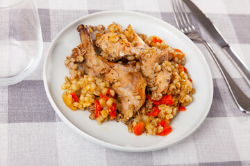 Portions of baked rabbit with porridge pearl barley are served on plate and supplemented with glass, knife, fork