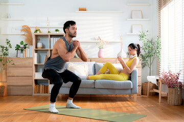 Obraz premium Young asian man doing squat exercise on yoga mat in living room at home while woman using phone for relax on sofa, fitness training and healthy lifestyle in a cozy, strength and strong of muscle.