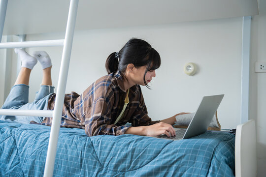 Student asian woman lying use laptop on bed in dorm room college student&nbsp;do homework on laptop in university dormitory.