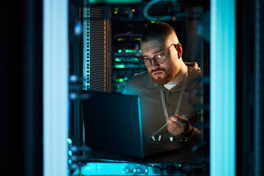 Man working in server room, analyzing data on laptop surrounded by racks of servers, maintaining focused expression throughout work setting part of technology maintenance - Powered by Adobe