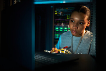 African American woman focused on maintaining server equipment in data center with various colored lights in background