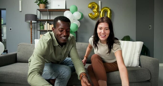 Happy multiracial and african american couple sitting on sofa, blowing in birthday trumpets at a bir