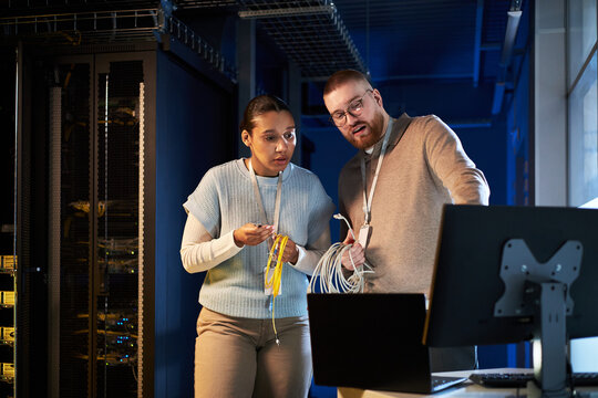 Caucasian female and Caucasian male engineers setting up network infrastructure in modern server room examining cables and hardware with serious expressions - Powered by Adobe