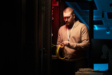 Technician in data center holding network cables, surrounded by server racks and computer equipment. Light illuminating from screens and devices, showcasing technological environment