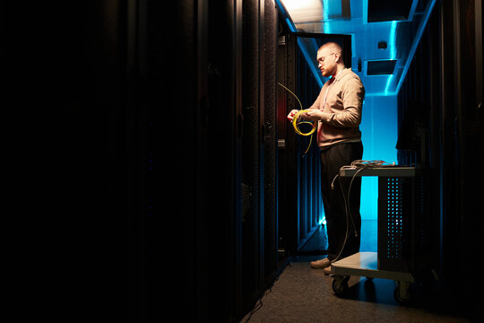 Technician working in data center, inspecting server racks and checking connections, lit by soft blue lights in a modern, secure facility
