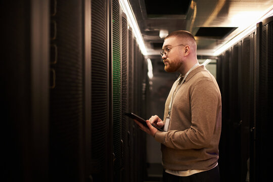 Individual monitoring servers in modern data center holding tablet in hand. Wearing glasses and observing various indicators on server racks - Powered by Adobe