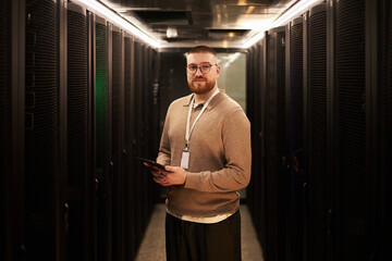 Portrait of young man with beard and glasses working in data center server room, standing between...