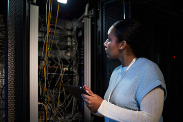 Woman holding tablet and examining server cables in a dimly lit server room. She appears focused on...