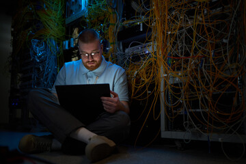 Person sitting on floor surrounded by numerous network cables in data center while working with...