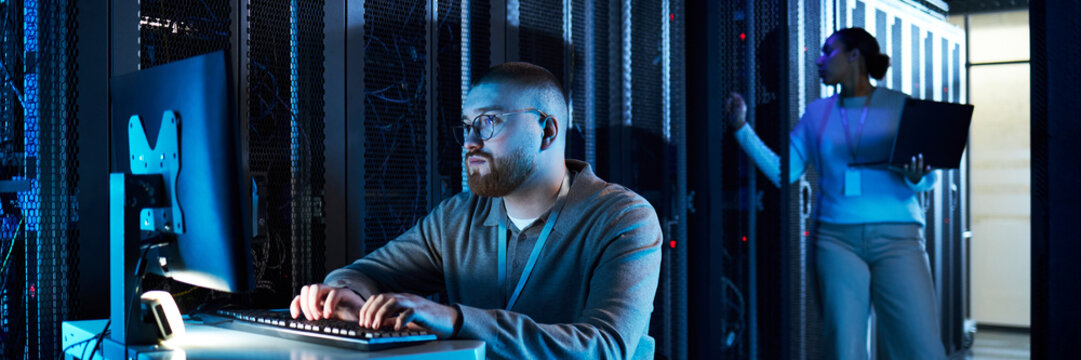 Male technician monitoring server activities on computer while female colleague holding laptop checking equipment in server room illuminated by blue lighting - Powered by Adobe