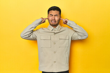 Asian man in stylish gray shirt on yellow studio covering ears with fingers, stressed and desperate by a loudly ambient.