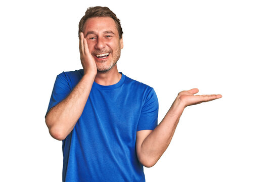 A middle-aged man isolated holds copy space on a palm, keep hand over cheek. Amazed and delighted. - Powered by Adobe