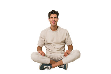 Young caucasian man sitting on the floor isolated on white background funny and friendly sticking out tongue.