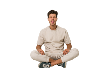 Young caucasian man sitting on the floor isolated on white background funny and friendly sticking out tongue.