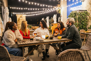 Naklejka premium Multiethnic group of friends enjoying lively drinks and engaging conversation at a trendy rooftop bar under the twinkling night sky