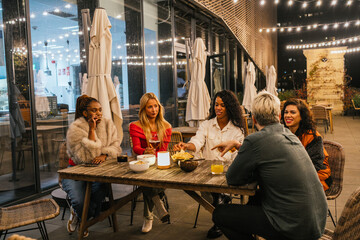 Naklejka premium Multiethnic group of friends having fun, eating, and drinking at a rooftop restaurant at night