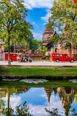 A scenic view of Chiang Mai city moat reflects the ancient Wat Lok Moli temple under a bright blue sky, with red songthaews and a tuk-tuk adding a touch of local life.