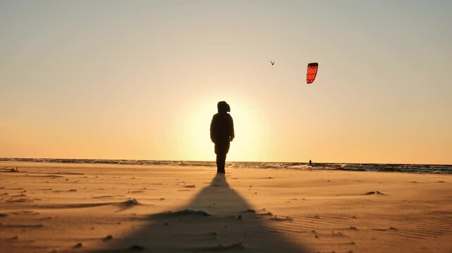 Lonely figure stands on Baltic coast as kite surfer sails under warm sunset sky