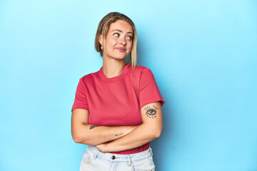 Blonde young woman in red t-shirt on blue background dreaming of achieving goals and purposes