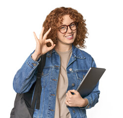 Young Caucasian redhead student with books cheerful and confident showing ok gesture.