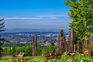 Totem poles in Burnaby Mountain Park on green grass next to trees and flower bed against blue sky,...